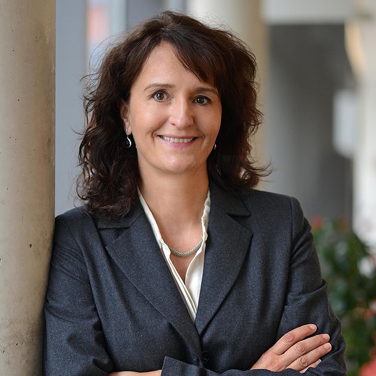 Eine Frau mit lockigem, braunem Haar steht lächelnd an einem Säulen-Element. Sie trägt einen grauen Blazer und eine helle Bluse. Ihr Arm ist verschränkt, und sie wirkt selbstbewusst und freundlich. Hintergrund unscharf, möglicherweise mit Pflanzen.