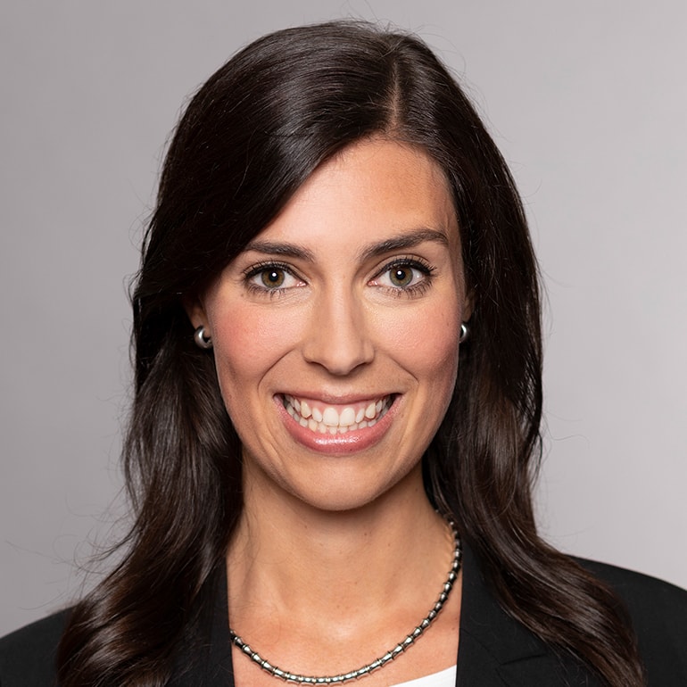 Das Bild zeigt eine lächelnde Frau mit langen, dunklen Haaren. Sie trägt ein schwarzes Blazer und eine elegante Kette. Der Hintergrund ist neutral und hell. Ihr Ausdruck vermittelt Freundlichkeit und Professionalität.