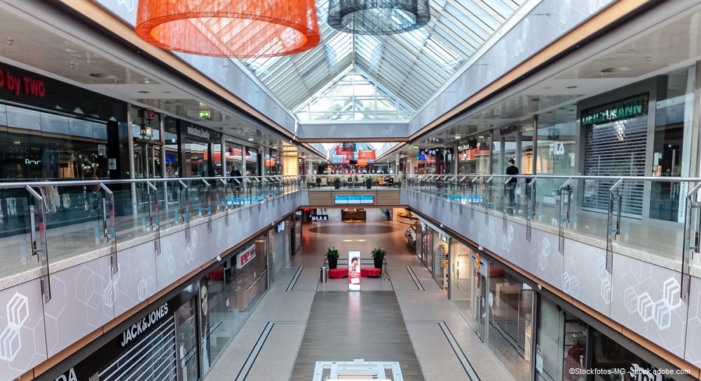 An indoor shopping mall features two levels, lined with retail stores along the walls. The spacious area has a glass roof allowing natural light in, and modern hanging lights add ambiance. People can be seen walking throughout the mall, creating a lively atmosphere.