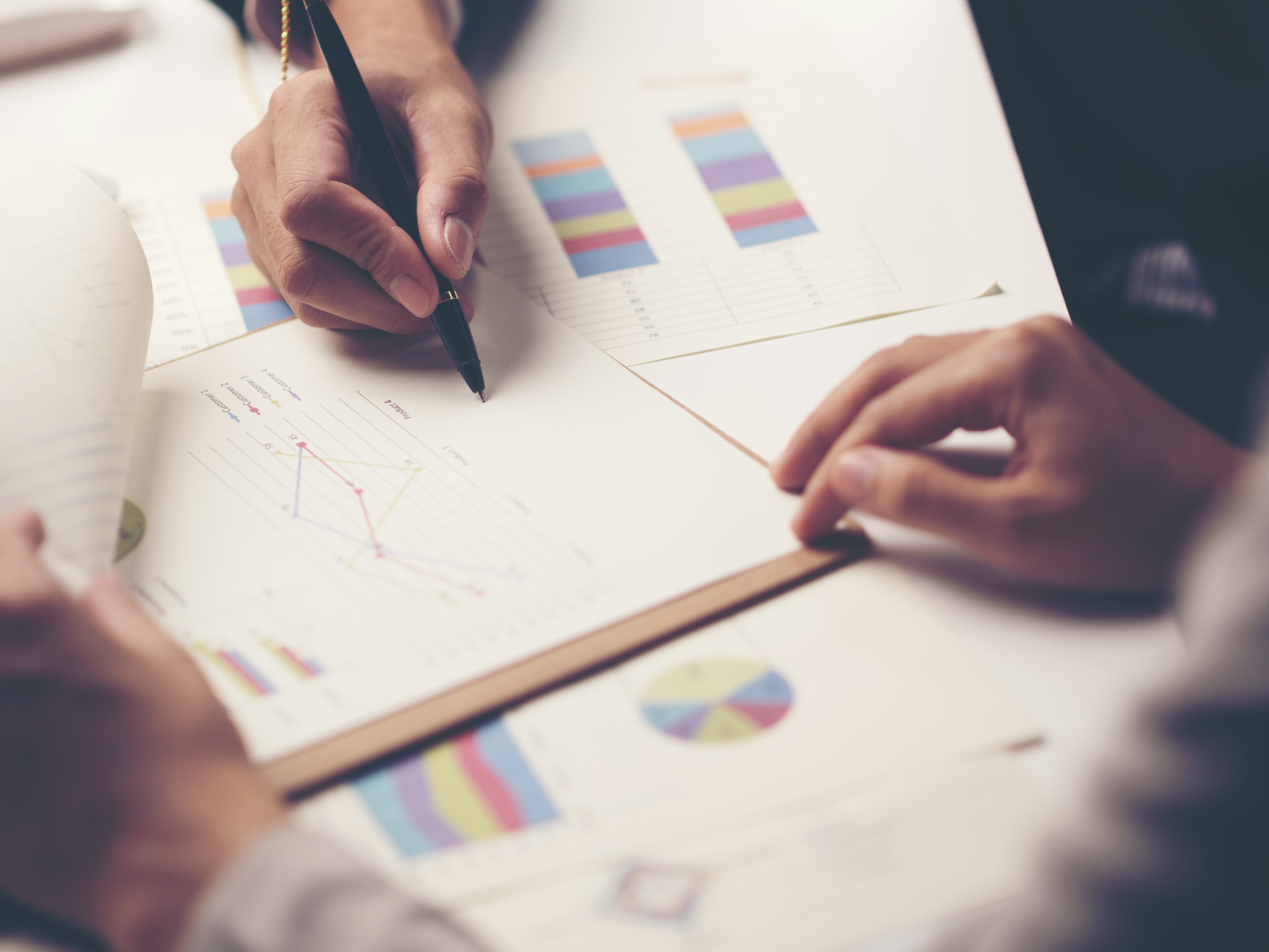 Two hands are engaged in a discussion over various documents that feature charts and graphs. One hand is holding a pen, while the other rests on the table. The papers show colorful data representations, indicating analysis or planning activities.
