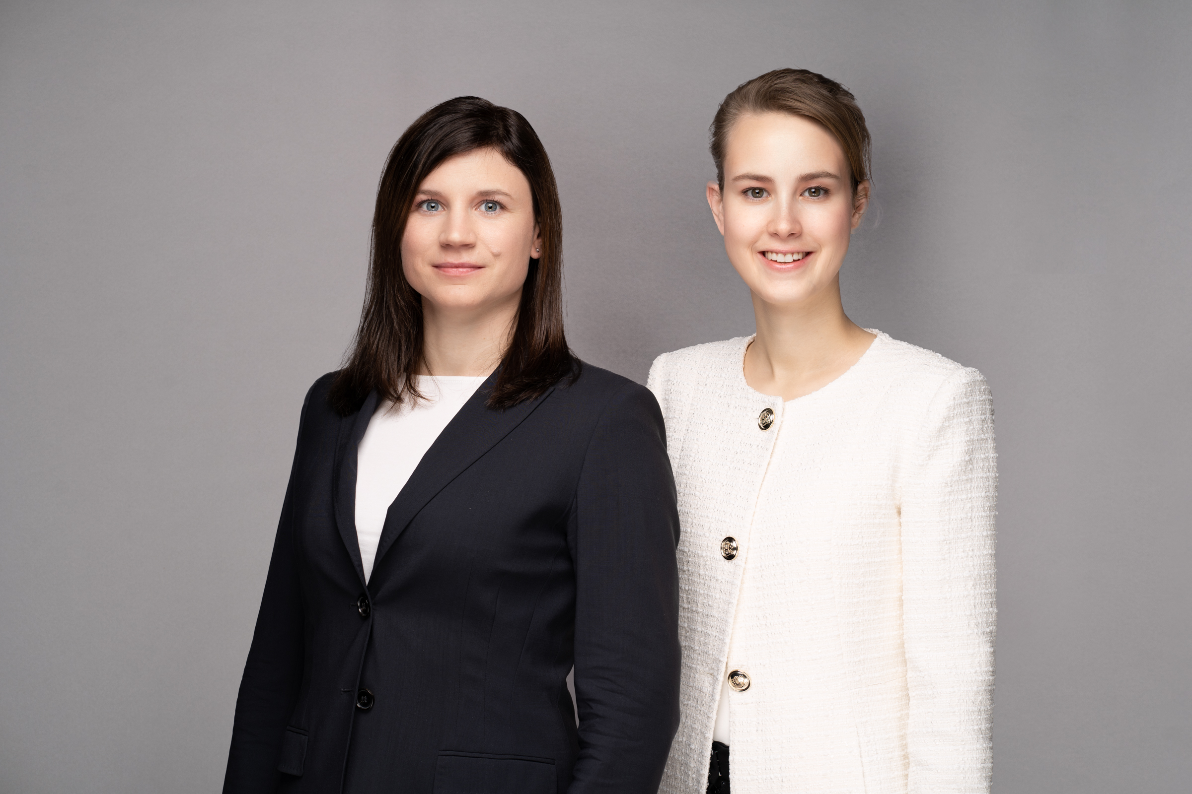 Zwei Frauen stehen nebeneinander vor einem grauen Hintergrund. Die Frau links trägt einen schwarzen Blazer und hat längeres, glattes, braunes Haar. Die Frau rechts trägt eine weiße Jacke und hat helles, kurzes Haar. Beide lächeln freundlich in die Kamerarichtung.