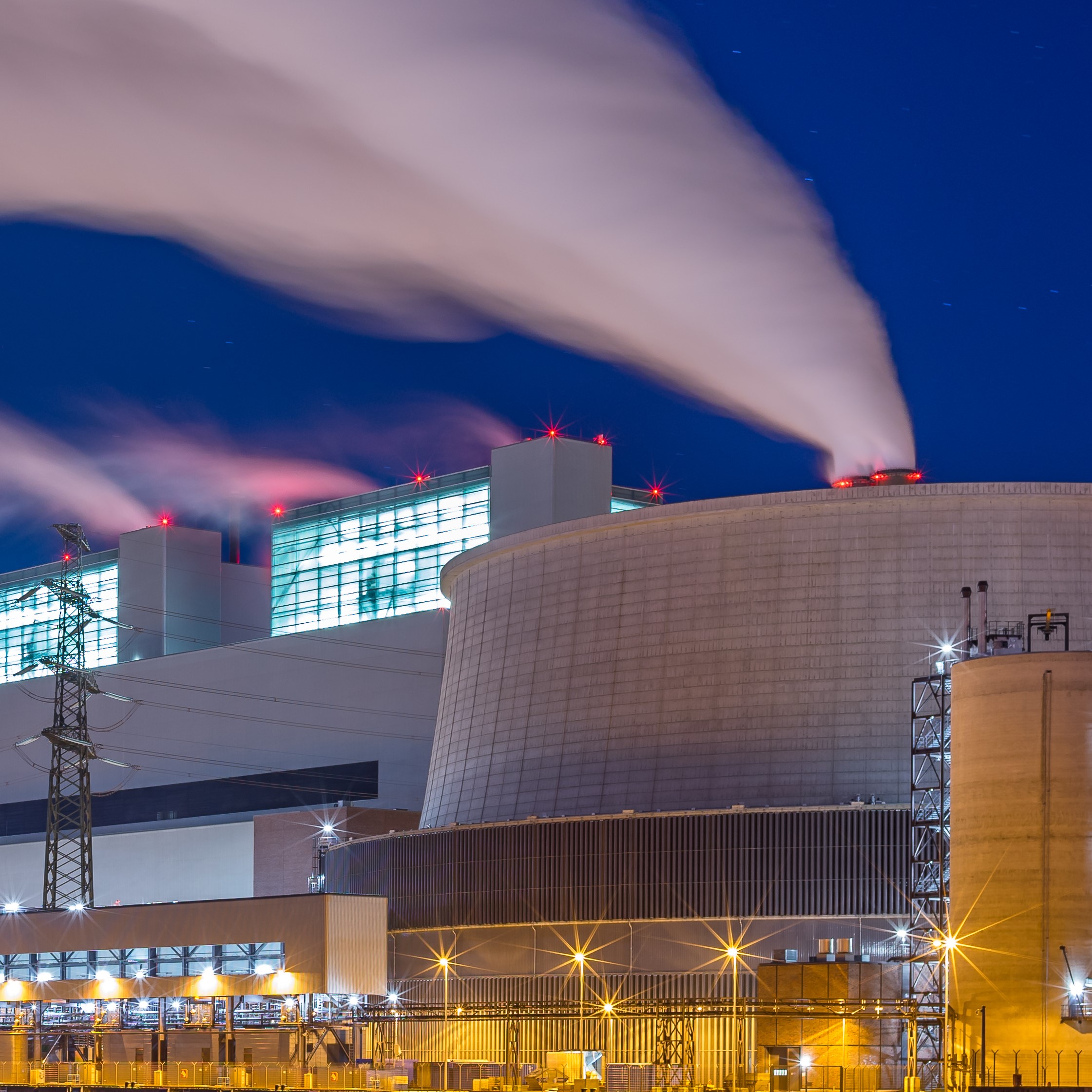 Ein modernes Kraftwerk bei Nacht mit sichtbaren Kühltürmen. Aus den Schornsteinen steigt Dampfwolken auf, während die Bürogebäude und die Umgebung beleuchtet sind. Die Architektur zeigt industrielle Elemente und eine klare Struktur.