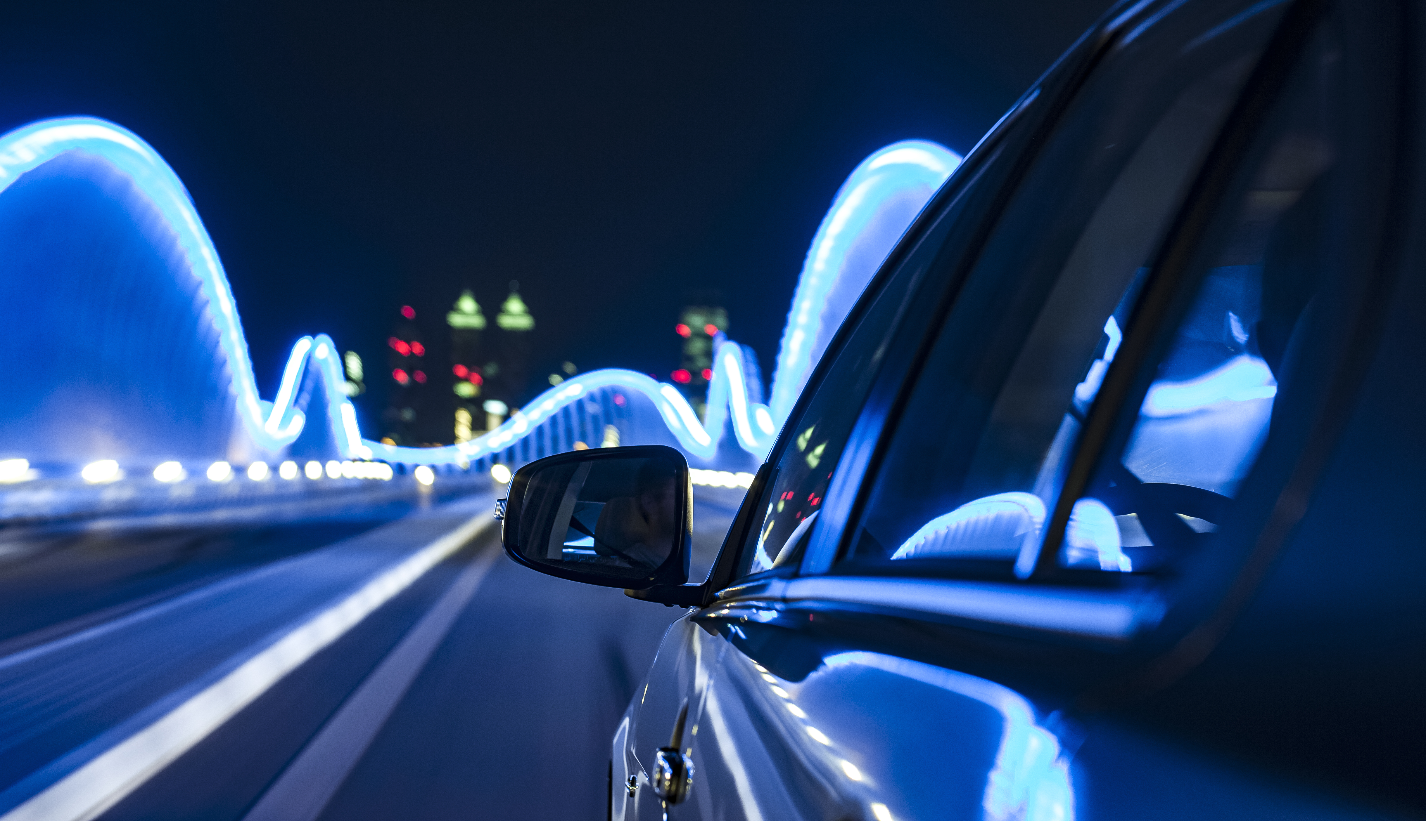 Ein Auto fährt schnell über eine beleuchtete Brücke bei Nacht. Die Umgebung ist von blauen Lichtern umgeben, während eine Stadtansicht im Hintergrund zu sehen ist. Der Fokus liegt auf dem Seitenspiegel des Fahrzeugs und der Bewegung der Straße.