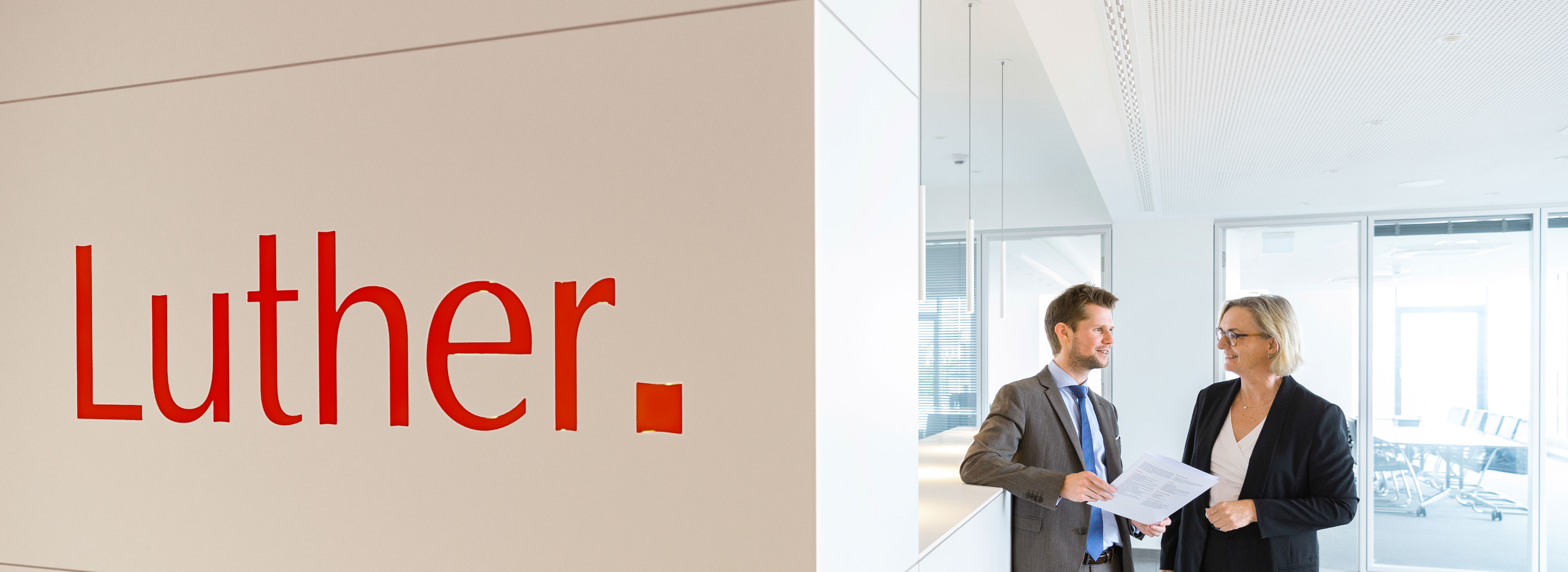 A man in a suit and a woman in a blazer are engaged in a conversation while holding documents. The background features a modern office space, and a prominent sign displays the name "Luther" in bold red letters.