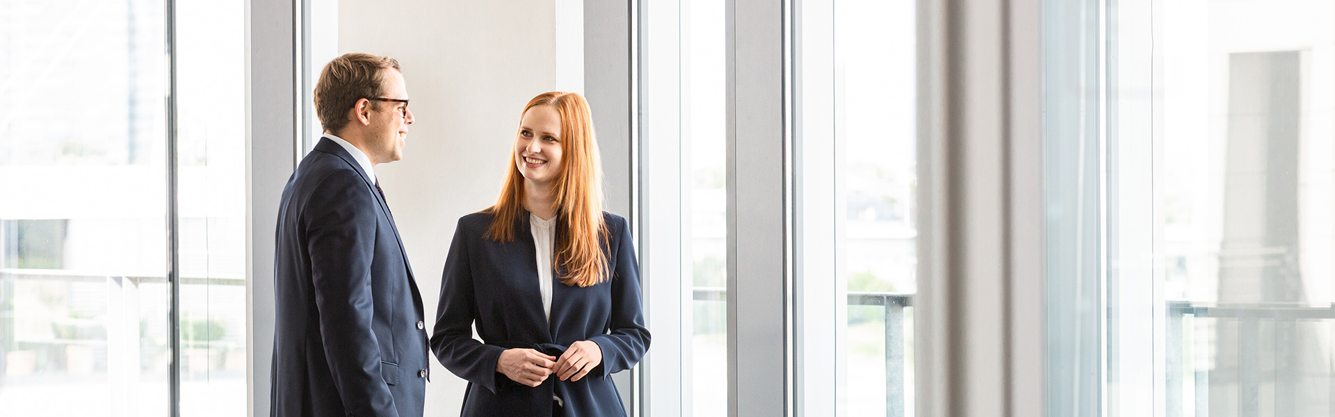 Two professionals engage in a conversation by large windows. The man is wearing glasses and a dark suit, while the woman, smiling, has long red hair and is dressed in a dark blazer. They share a friendly, collaborative demeanor in a bright, modern space.