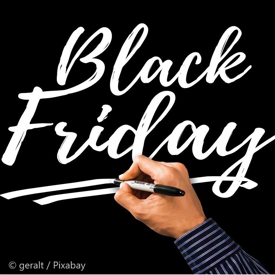 A hand holding a black marker is writing "Black Friday" in bold, white lettering against a black background. The text is slightly slanted and includes an underlined section below the words.