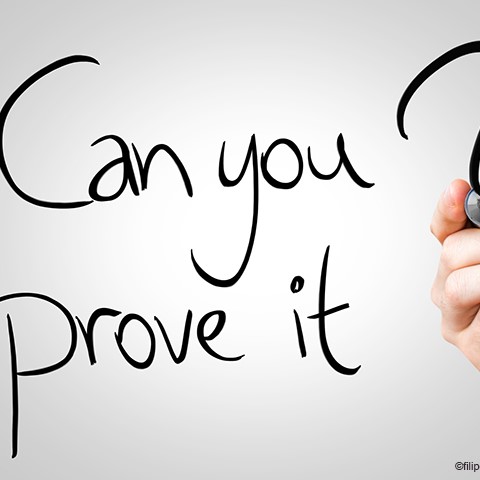 Ein Hand, die einen Stift hält, schreibt in klarer Schrift: "Can you prove it?" Der Hintergrund ist schlicht und neutral, was den Fokus auf den Text lenkt.