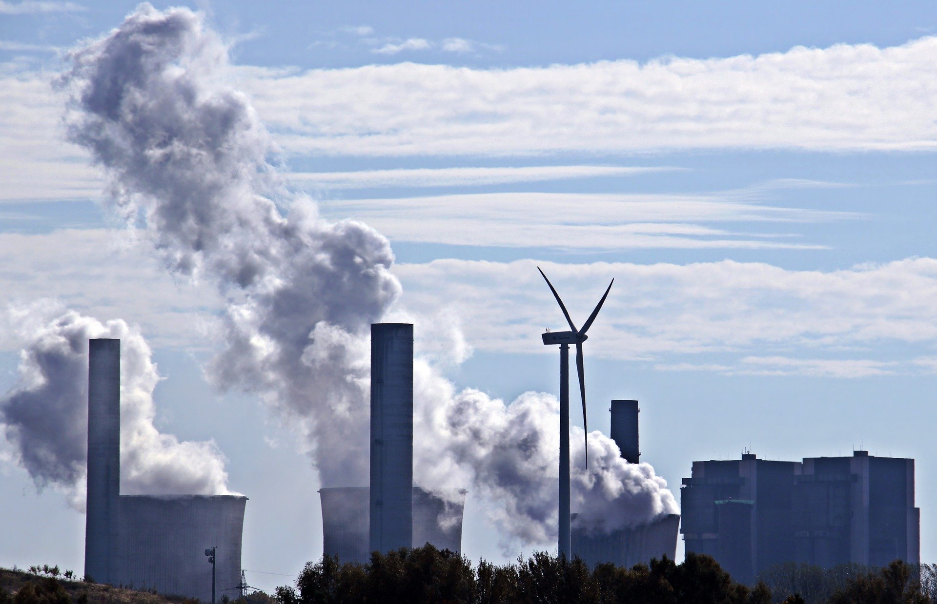 The image depicts industrial smokestacks releasing thick white smoke against a blue sky. In the foreground, a wind turbine stands as a contrast to the pollution, symbolizing renewable energy alongside traditional fossil fuel power generation.