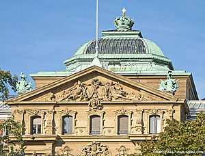 The image depicts the ornate upper section of a historic building featuring a green copper dome with a central skylight. The facade is richly decorated with sculptures and reliefs, showcasing intricate architectural details against a clear blue sky.