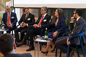 Eine Podiumsdiskussion mit fünf Personen, die auf Stühlen sitzen. Eine Frau hält ein Mikrofon und spricht, während die anderen aufmerksam zuhören. Im Hintergrund ist eine Leinwand und einige Pflanzen zu sehen. Tische mit Wasserflaschen sind ebenfalls vorhanden.