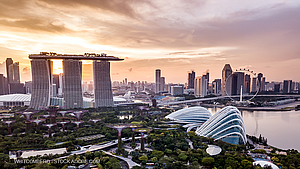 A panoramic view of Singapore showcases the iconic Marina Bay Sands hotel alongside the futuristic Gardens by the Bay domes. In the distance, the Singapore Flyer, a large observation wheel, is visible against a vibrant sunset sky, reflecting the city's blend of modern architecture and nature.