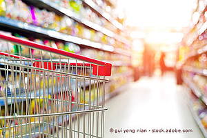 A shopping cart is positioned in the foreground of a grocery aisle filled with colorful products on the shelves. The background is slightly blurred, suggesting movement and activity, possibly indicating shoppers in the distance.