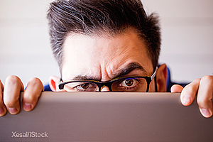 A man with glasses is peering over the edge of a laptop, his brow furrowed and eyes focused, showing a look of concern or concentration. The background is blurred, emphasizing his expression and the device in front of him.