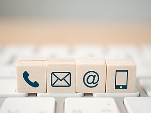 Vier Holzblöcke mit Symbolen für Kontaktmöglichkeiten: ein Telefonhörer, ein Briefumschlag für E-Mail, das @-Symbol und ein Smartphone. Die Blöcke sind auf einer Tastatur platziert, um verschiedene Kommunikationswege darzustellen.