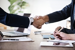 Two people are shaking hands over a wooden table, signifying a business agreement or partnership. A laptop and various papers are visible on the table, along with a person writing notes.