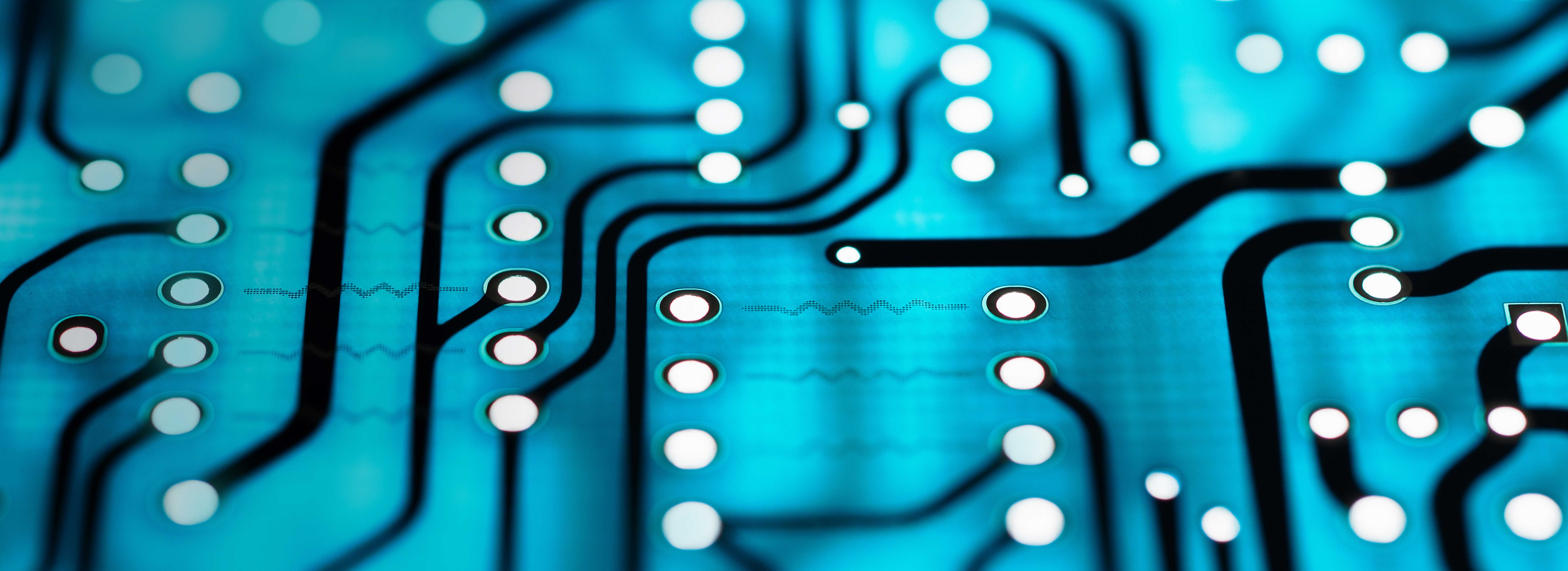 Nahaufnahme einer Leiterplatte mit blauen und schwarzen Leiterbahnen sowie runden Kontaktpunkten. Die feinen Linien und Muster verdeutlichen die Komplexität elektronischer Schaltungen. Der Fokus liegt auf der Struktur und den Verbindungen, die in modernen elektronischen Geräten verwendet werden.