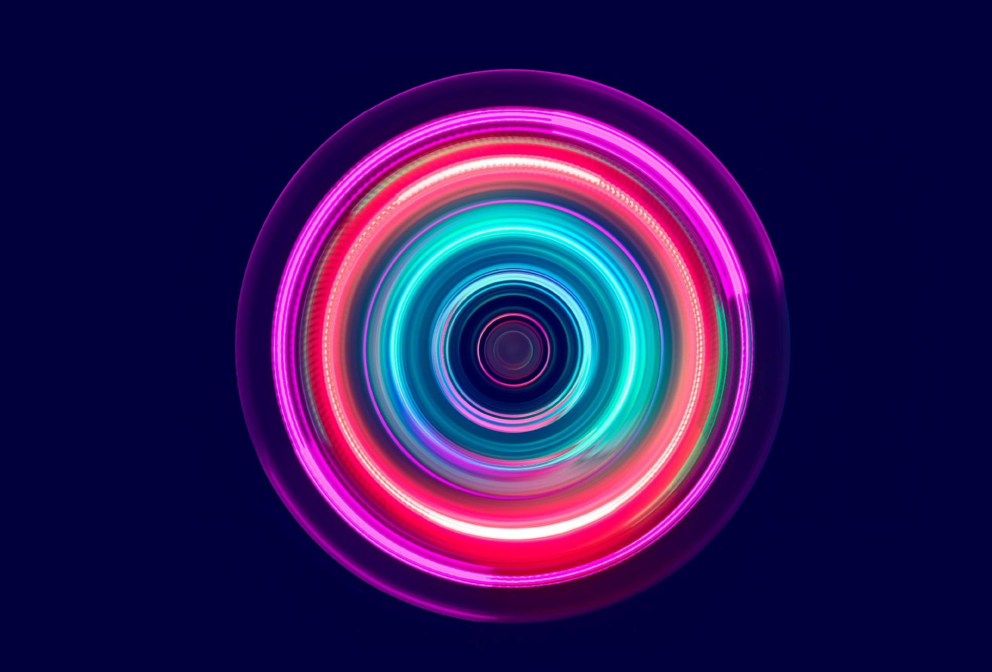 Ein kreisförmiges Muster aus leuchtenden Farben in Blau, Pink und Grün. Die Farben scheinen sich zu bewegen und erzeugen einen hypnotisierenden Effekt. Der Hintergrund ist dunkel, wodurch die Lichtstrahlen hervorstechen.