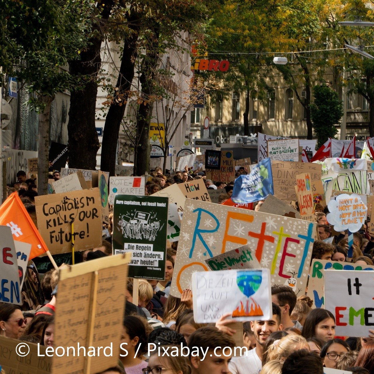 A large crowd of protesters holds colorful signs advocating for climate action and social justice. Messages include "Capitalism Kills Our Future" and "Save the Planet." The scene indicates a strong public demonstration focused on urgent environmental and societal issues.