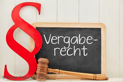 Ein rotes Paragraphenzeichen steht neben einer kleinen Tafel, auf der in weißer Schrift "Vergaberecht" geschrieben steht. Darunter liegt ein Richterhammer. Die Tafel ist aus Holz, und der Hintergrund ist hell und einfarbig.