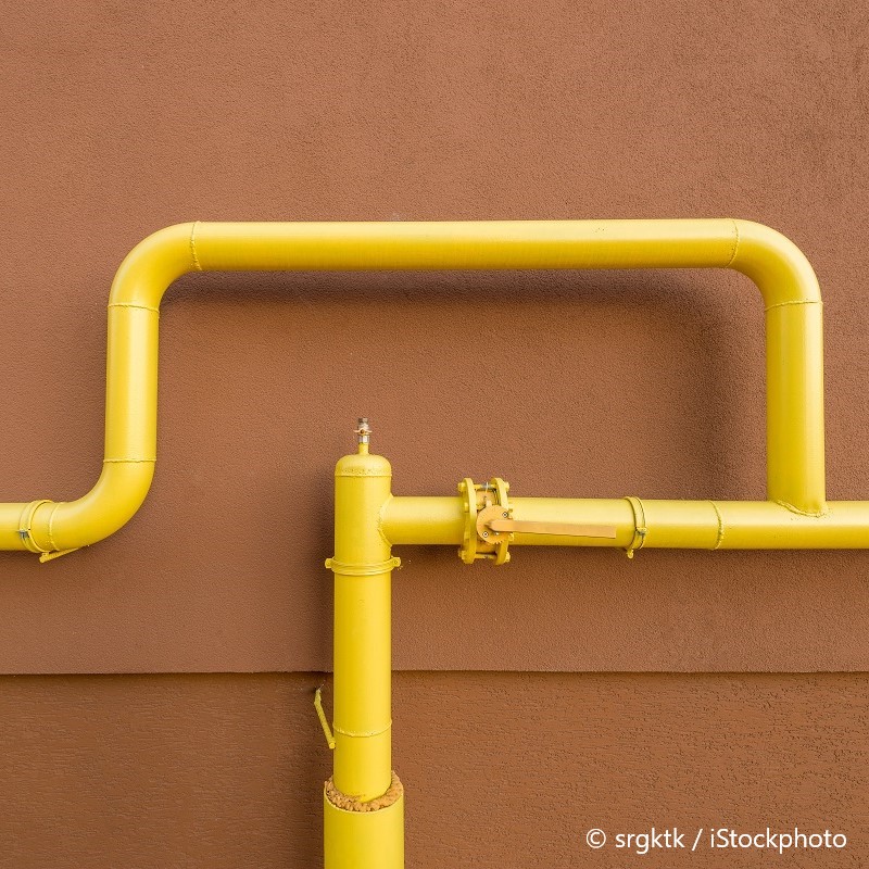 A close-up of a yellow pipe system attached to a brown wall. The pipes are arranged in an angular pattern, with various fittings and a valve visible. The bright yellow color contrasts sharply with the muted brown background, emphasizing the design and structure of the piping.