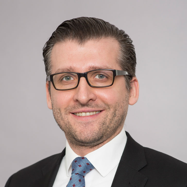 Ein lächelnder Mann mit Brille, schwarzem Anzug und weißem Hemd, trägt eine geblümte Krawatte. Der Hintergrund ist einfarbig grau. Die Darstellung vermittelt ein freundliches und professionelles Auftreten.