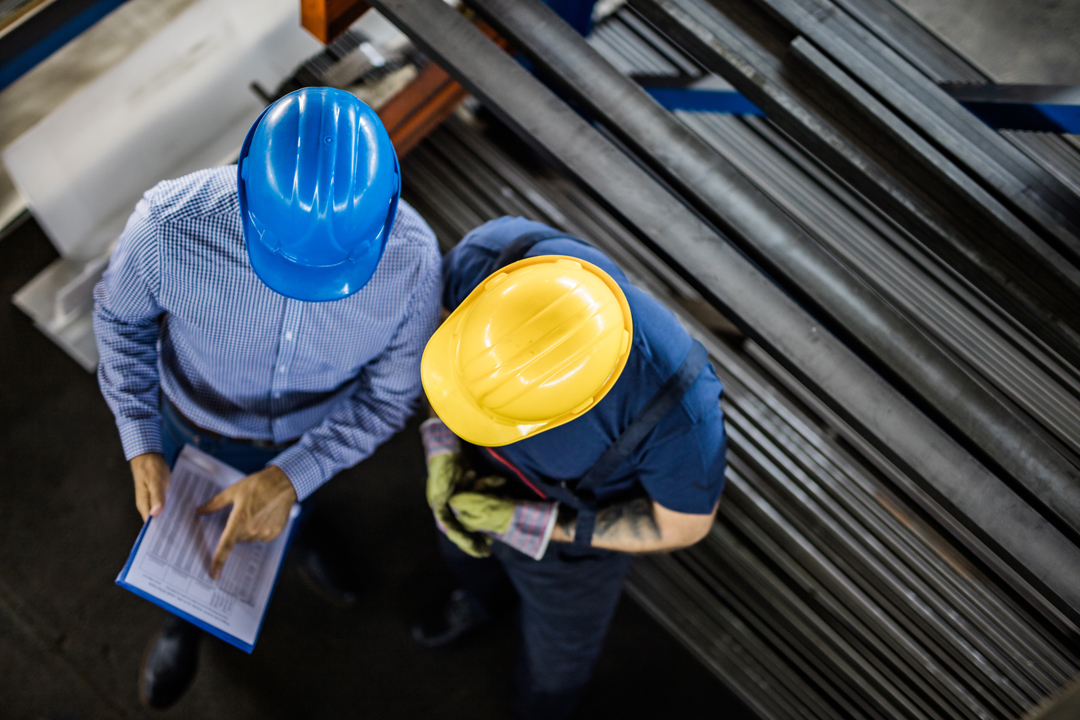 Two individuals in safety helmets are discussing work-related documents in an industrial setting. One person wears a blue helmet and the other a yellow helmet, while standing near metallic materials. The environment indicates a workshop or factory atmosphere.