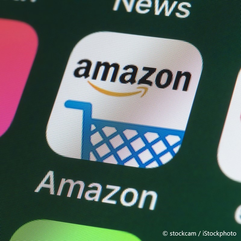 The image displays the Amazon app icon, featuring the word "Amazon" in black text alongside a yellow arrow and a blue shopping cart graphic. The background is white, and the icon is surrounded by other app icons.