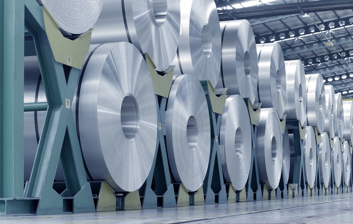 A series of large metallic rolls of aluminum are stacked neatly in a warehouse. The rolls are arranged on green stands, showcasing their shiny, smooth surfaces and circular shapes. The setting appears industrial, with a high ceiling and structured lighting.