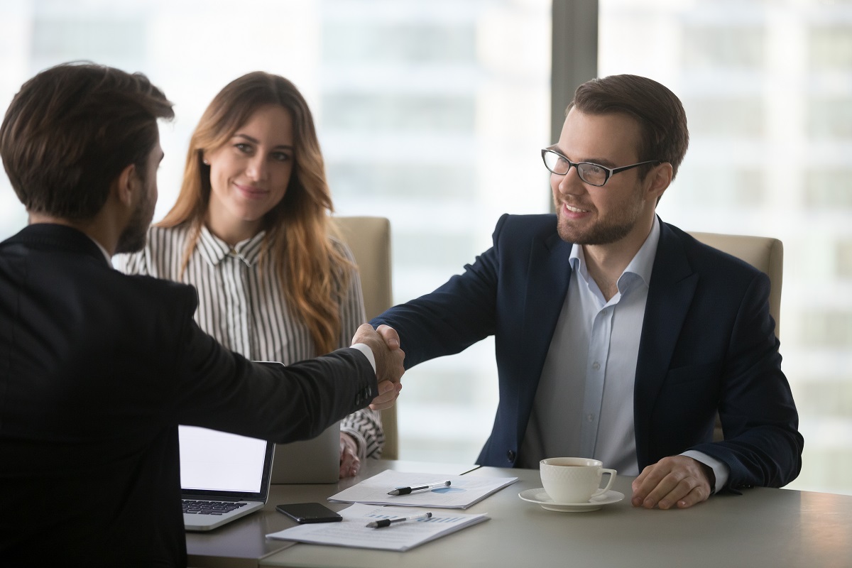 A business meeting setting features two men shaking hands in a friendly gesture. A woman sits in the background, smiling. The table is organized with a laptop and documents, suggesting a collaborative discussion. Natural light filters in, creating a professional atmosphere.