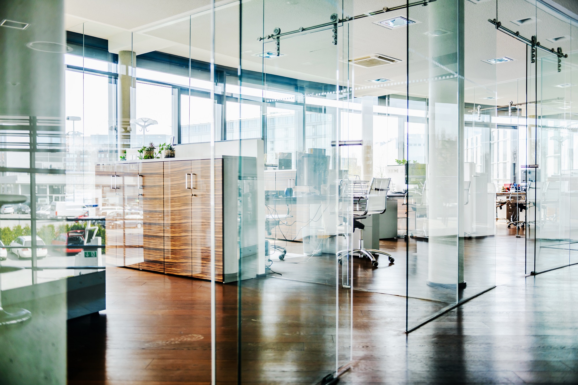 A modern office space featuring large glass walls and wooden flooring. In the foreground, there's a wooden storage cabinet and potted plants. Behind, office desks and chairs are visible, with large windows allowing natural light and a view of the outside.