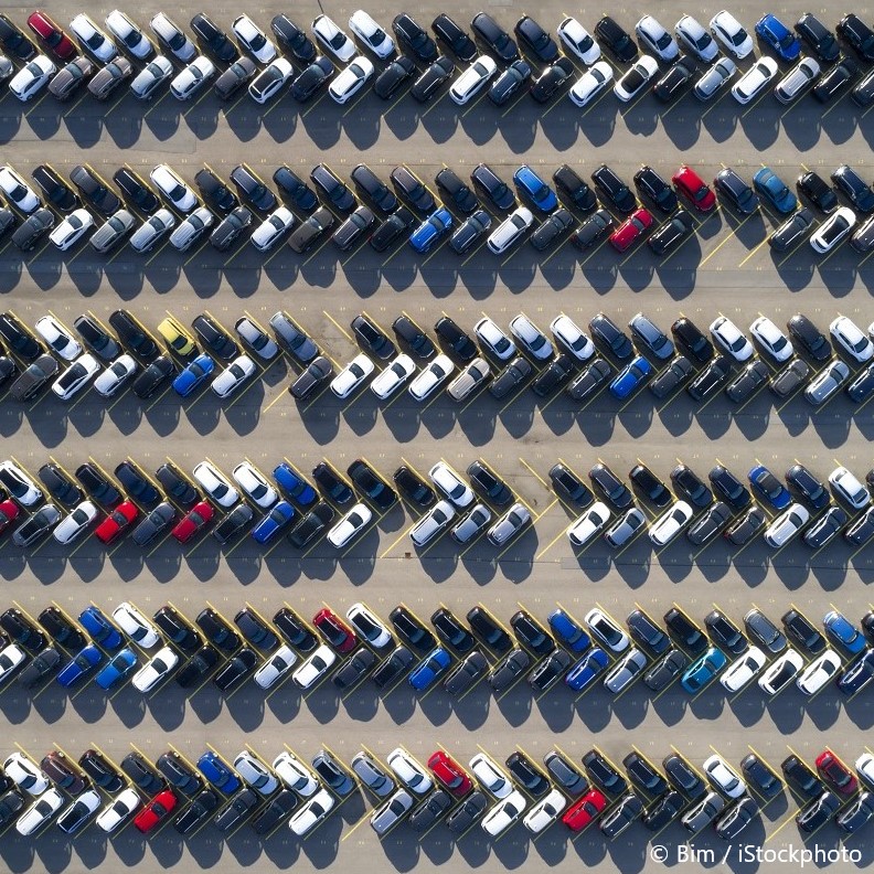 An aerial view of a parking lot filled with vehicles, arranged in rows. The cars are of various colors, including white, black, blue, and red, all parked close together in an organized pattern, showcasing a dense arrangement of automobiles.