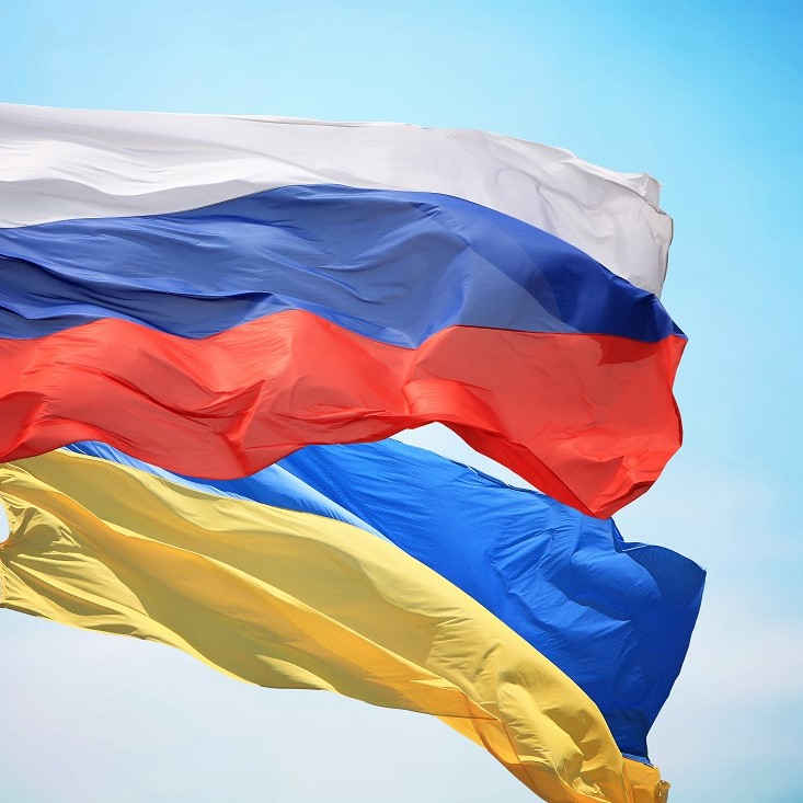 Two flags are prominently displayed: the flag of Russia, featuring horizontal stripes of white, blue, and red, above the flag of Ukraine, which has horizontal stripes of blue and yellow. The flags are waving against a clear blue sky.