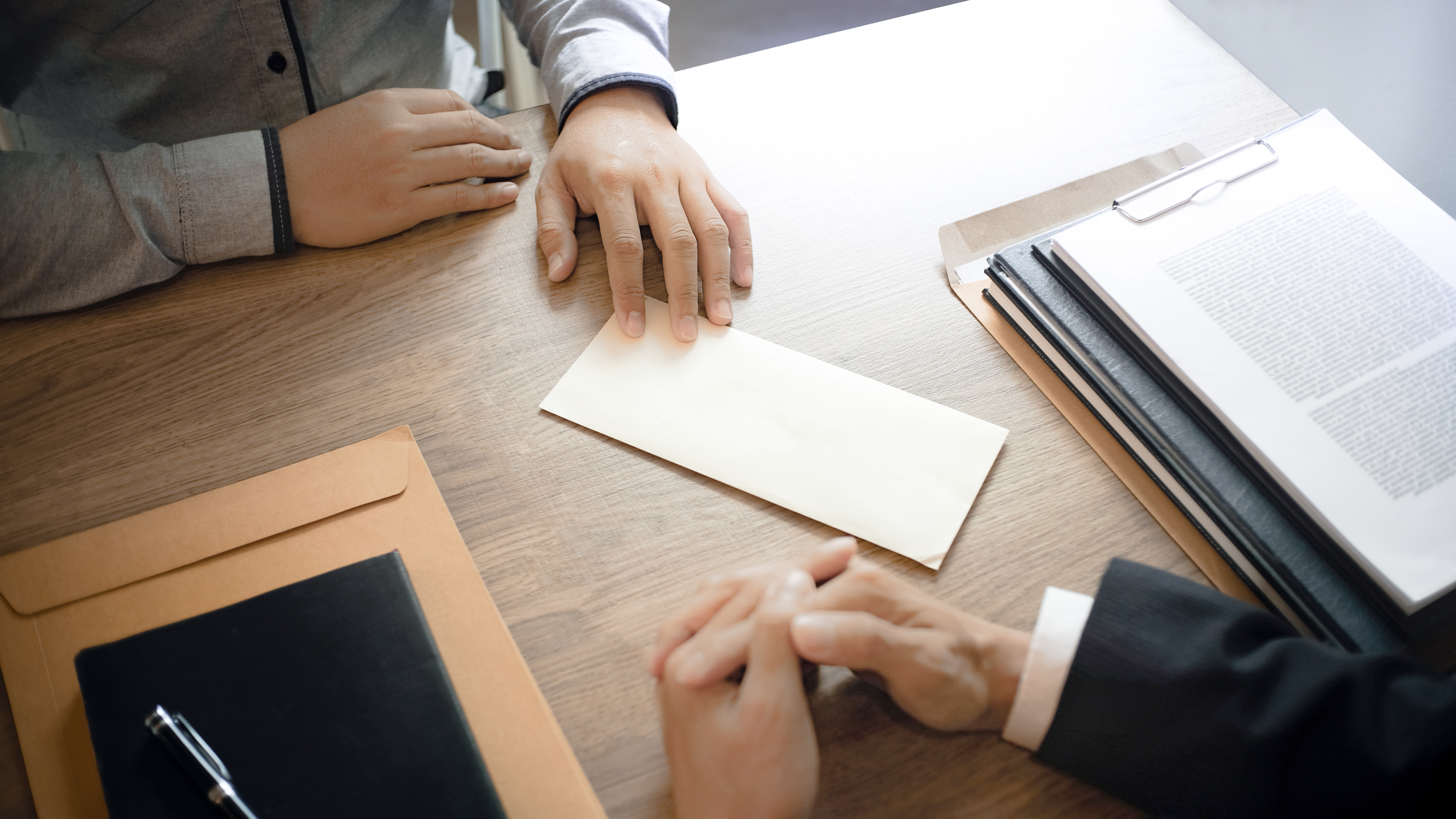 Two people are seated at a wooden table. One person, wearing a gray shirt, has their hand resting near a blank envelope, while the other, dressed in a suit, clasps their hands together. Documents are stacked on one side of the table.
