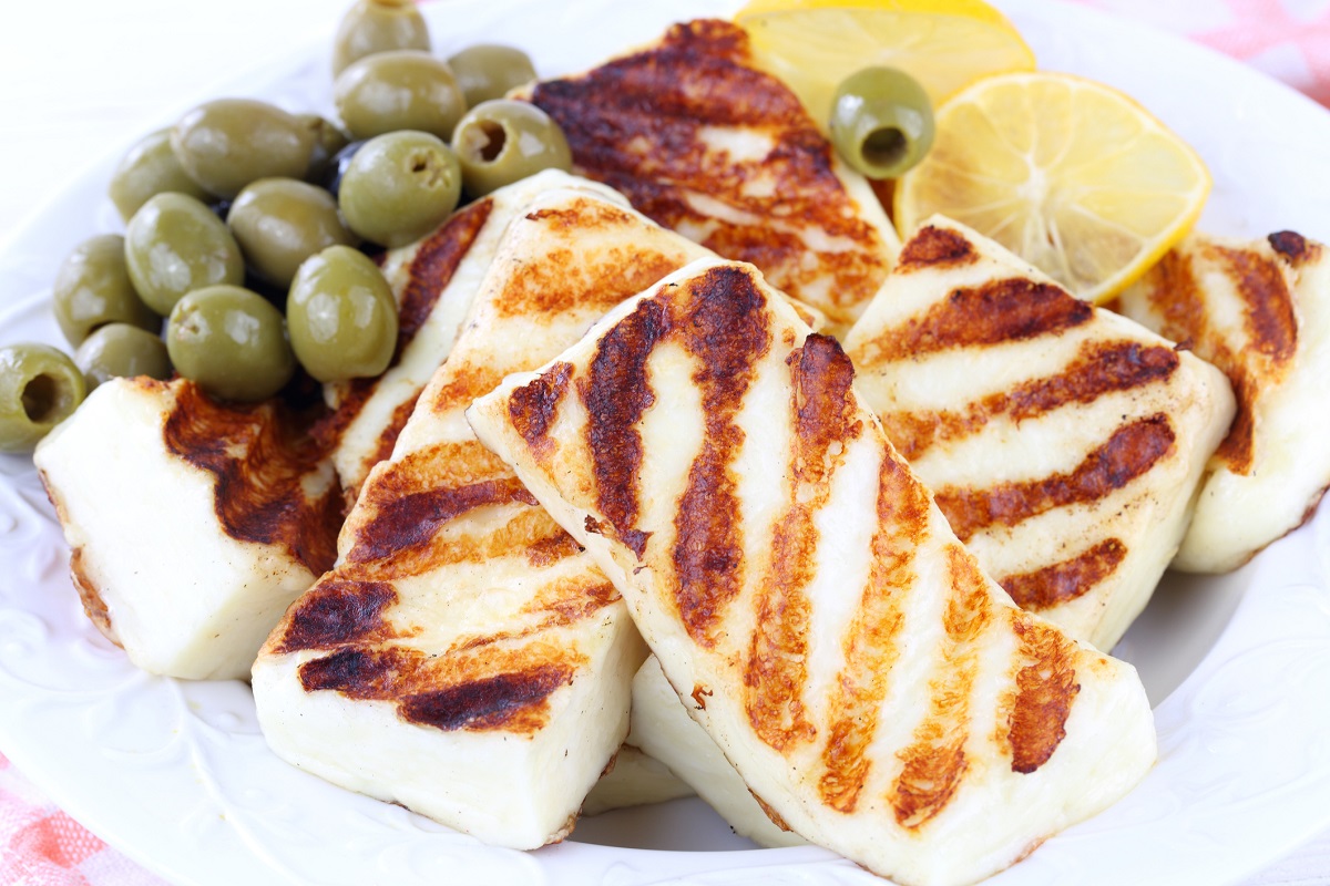 A plate of grilled halloumi cheese slices with distinct grill marks, accompanied by green olives and lemon wedges. The dish showcases a combination of textures and colors, inviting a flavorful experience.