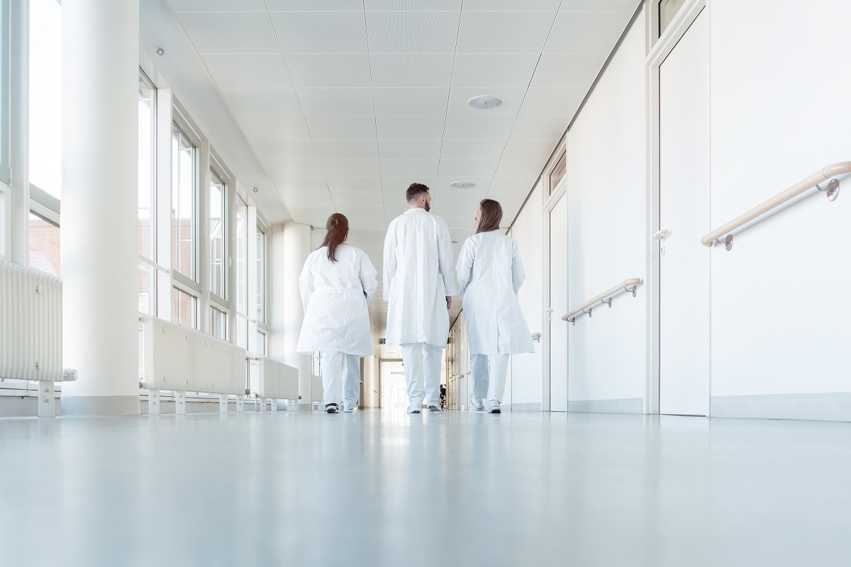 Drei medizinische Fachkräfte in weißen Kitteln gehen einen hellen Krankenhausflur entlang. Der Flur ist lichtdurchflutet und modern gestaltet. Die Personen sind von hinten zu sehen und scheinen sich auf eine Besprechung oder einen Einsatz zuzubewegen.