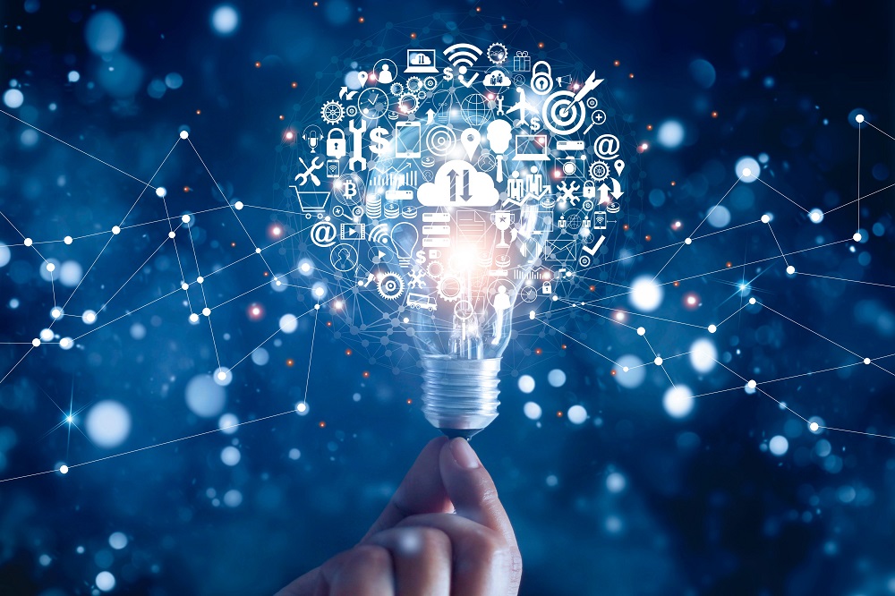 Eine Hand hält eine Glühbirne, aus der zahlreiche Symbole und Icons strömen, die Konzepte wie Ideen, Technologie und Vernetzung darstellen. Der Hintergrund ist dunkel mit leuchtenden Punkten, die eine dynamische Atmosphäre schaffen.