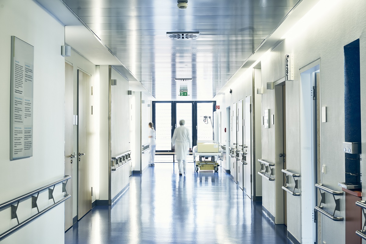 A hospital corridor is shown, featuring white walls and shiny blue floors. Medical staff are seen walking, and patient rooms line the sides. Large windows at the end of the hallway allow natural light to enter. A bed on wheels is positioned nearby, indicating a medical setting.
