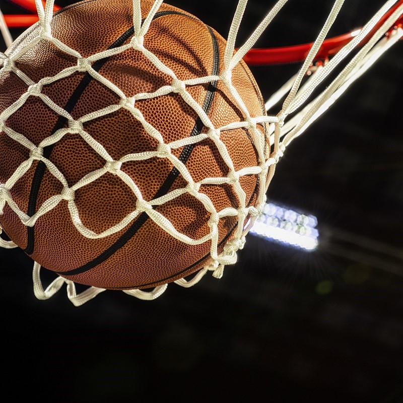 A close-up view of a basketball caught in a white net, suspended beneath a basketball hoop. The ball's texture is visible, highlighting its surface and the white net's intricate pattern around it. The background is dark, emphasizing the basketball and net.