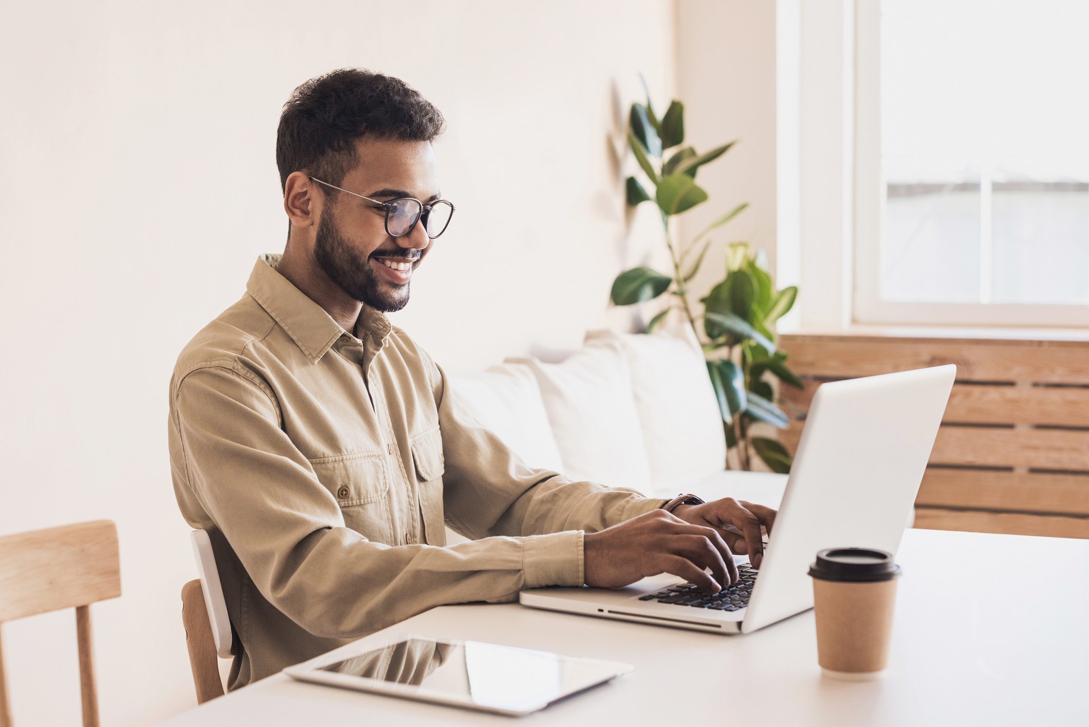 A smiling young man with glasses sits at a table, typing on a laptop. He wears a light brown shirt, and a coffee cup is placed beside him. In the background, a plant and a light setting create a cozy atmosphere.