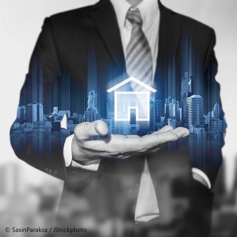 A businessperson in a suit holds an open hand displaying a glowing house icon. In the background, a city skyline is subtly visible, conveying a theme of real estate and urban development. The image uses a combination of monochrome and blue tones.
