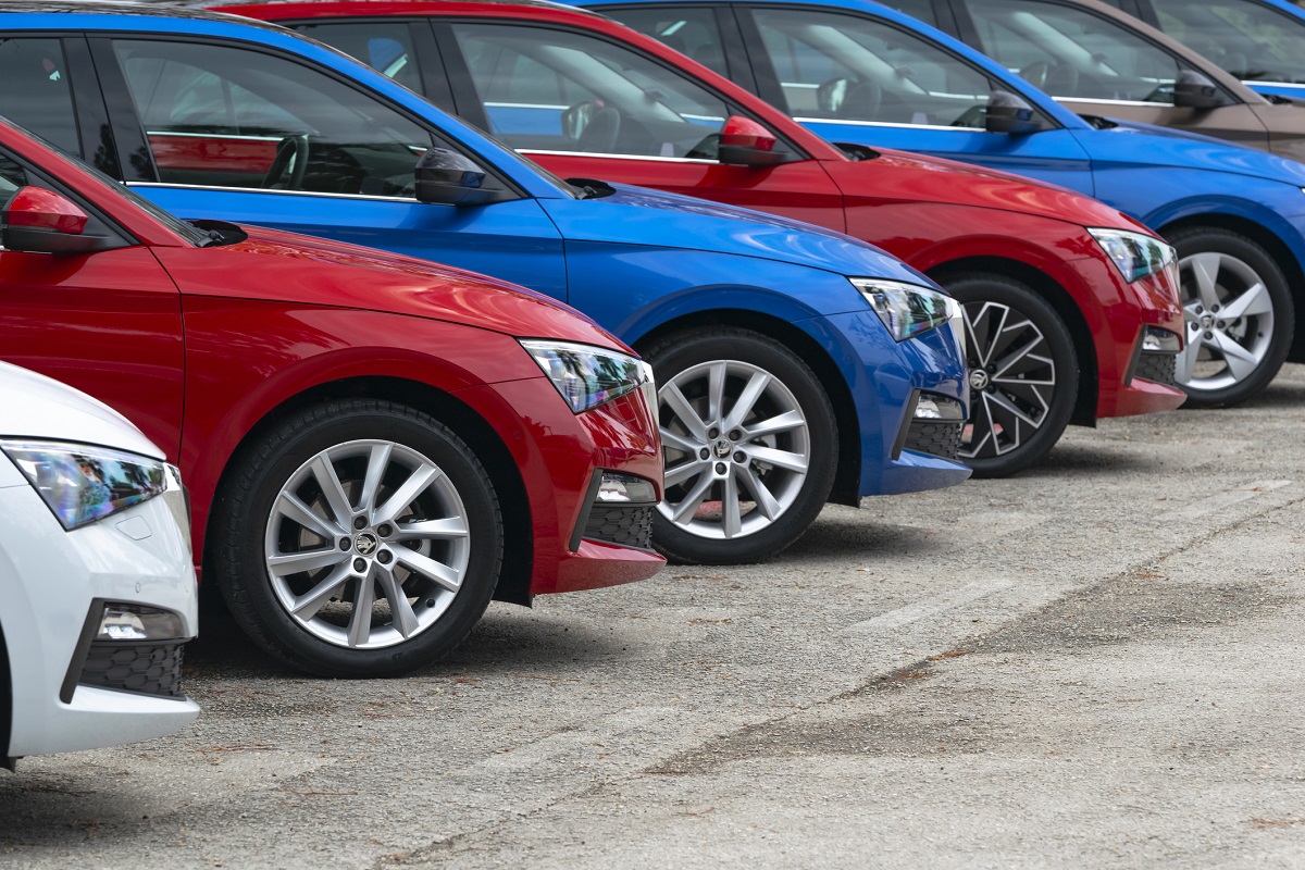 A row of parked cars in various colors, including white, red, and blue. The focus is on the side profiles, showcasing their wheels and sleek designs arranged neatly on a concrete surface.