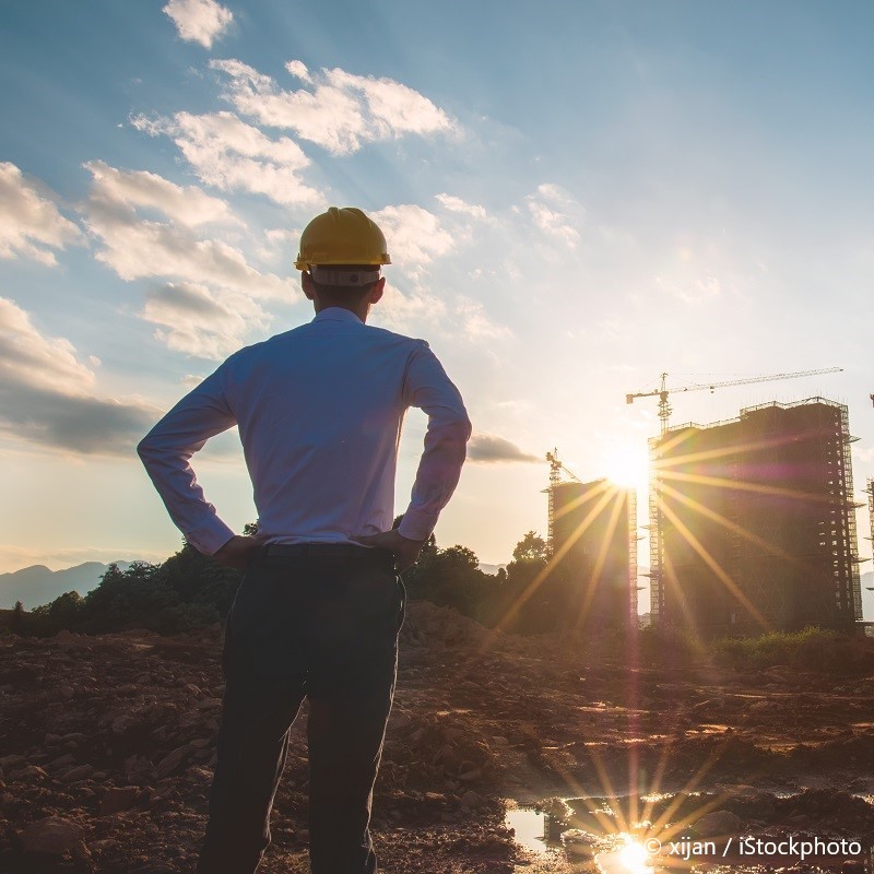 A construction worker in a hard hat stands with hands on hips, gazing at a building site under a sunset. The scene features cranes and construction structures in the background, with rays of sunlight illuminating the landscape.