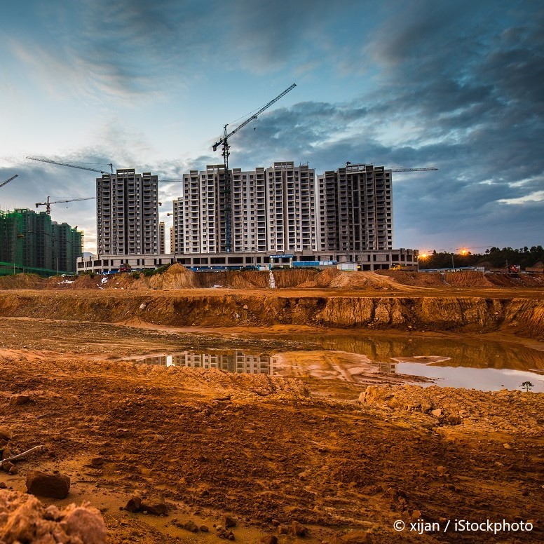 A construction site features several multi-story buildings in various stages of development. Cranes tower above, and a muddy landscape with earthworks and a small puddle is visible in the foreground. The sky is partially cloudy, indicating either dawn or dusk.