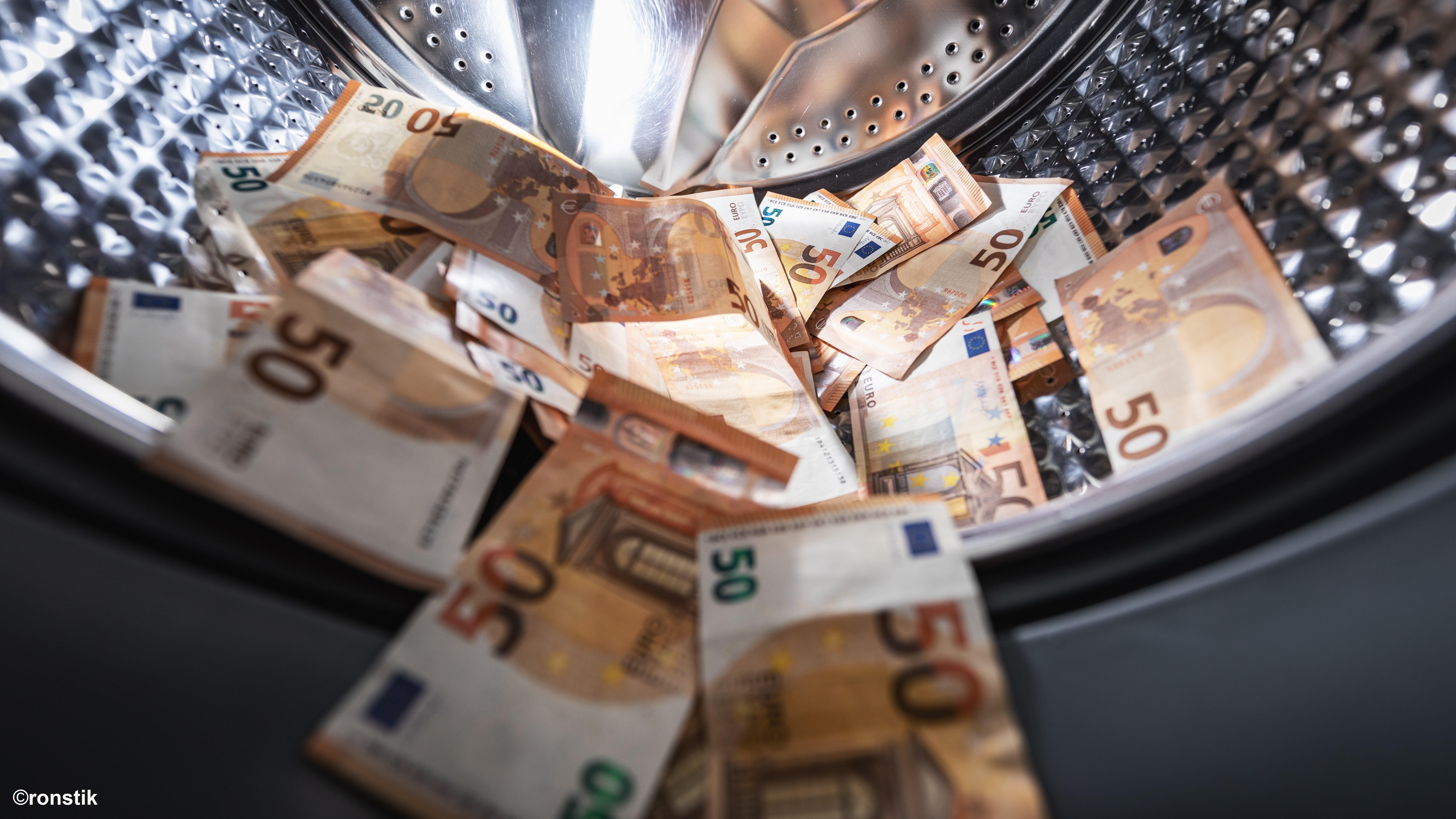 A close-up view inside a washing machine drum filled with various Euro banknotes. Predominantly seen are several crumpled 50 Euro notes, creating a chaotic yet colorful mix among the metallic interior of the washing machine.