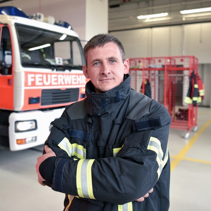 A confident firefighter in uniform stands with crossed arms, smiling. Behind him, a fire truck is parked, displaying the word "FEUERWEHR." The background includes racks holding firefighting gear, emphasizing the fire station setting.