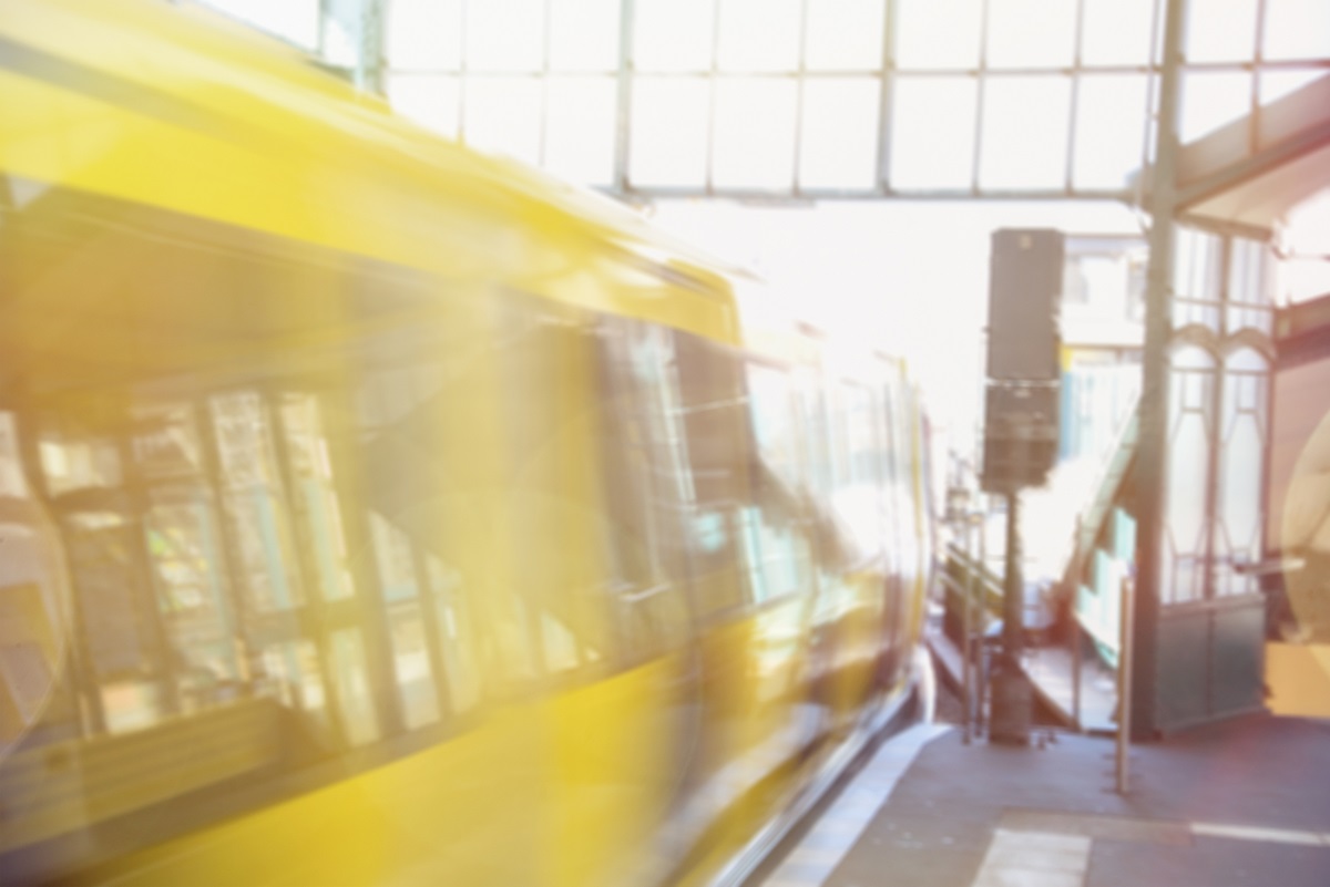 A speeding yellow train moves through a station, with sunlight filtering into the space. The platform is partially visible, suggesting an urban transit environment. The image conveys a sense of motion and energy associated with public transportation.
