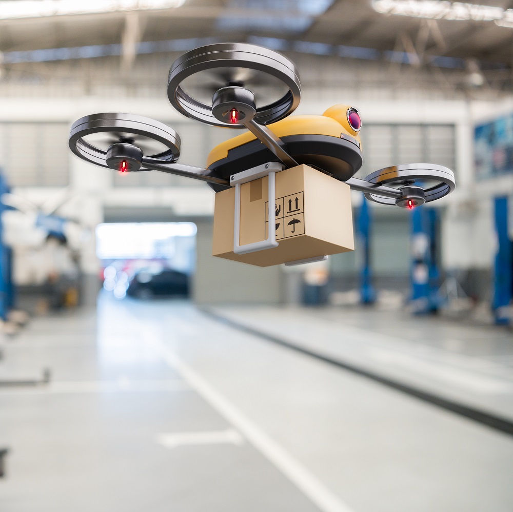 A yellow drone hovers in a modern warehouse, carrying a small package. The drone features four propellers, with red lights indicating its operational status. The background shows an industrial setting, emphasizing the technology used for delivery.