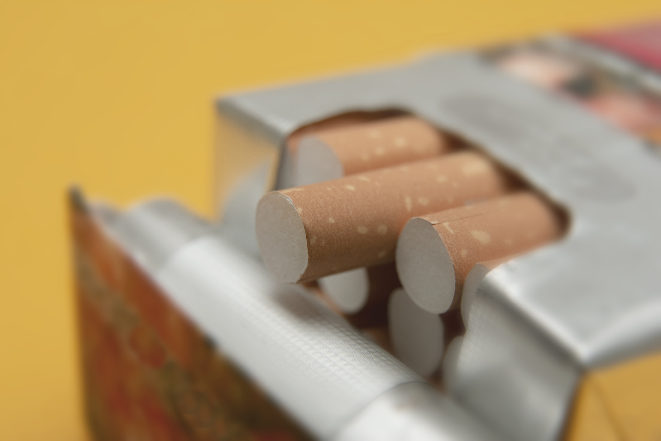 Eine geöffnete Zigarettenschachtel, aus der mehrere Zigaretten herausragen. Die Zigaretten haben eine braune Hülse mit einer weißen Spitze. Der Hintergrund ist gelb, was einen starken Kontrast zur Schachtel und den Zigaretten bildet.