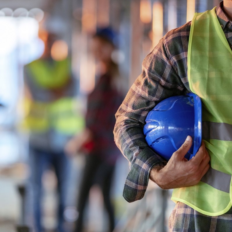 A construction worker in a green safety vest holds a blue hard hat close to his body. In the background, additional workers in safety vests are slightly blurred, indicating an active construction site. The setting suggests a collaborative work environment focused on safety.
