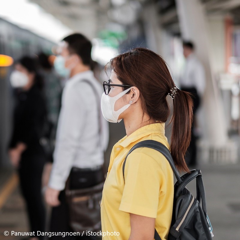 Eine Frau mit Brille und Mundschutz steht an einem Bahnhof. Sie trägt ein gelbes Shirt und hat einen Rucksack dabei. Im Hintergrund sind weitere Menschen zu sehen, die ebenfalls auf den Zug warten.